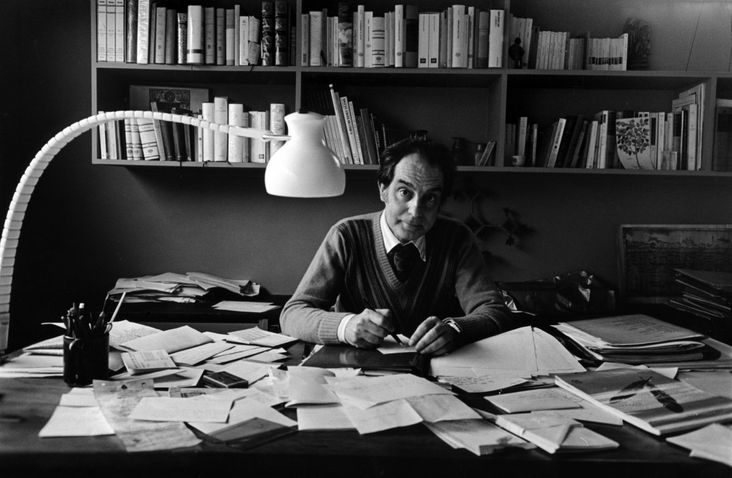 fotografia mostra Italo Calvino sentado em escrivaninha repleta de papeis, com estantes de livros na parede atrs